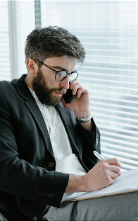 Businessman in glasses and black jacket taking notes during a phone call by a window with blinds