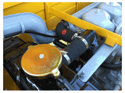 Zbliżenie na komorę silnika klasycznego samochodu, widoczny złoty korek chłodnicy, czarne gumowe przewody chłodzące z czerwonym logo oraz żółta belka wzmacniająca.