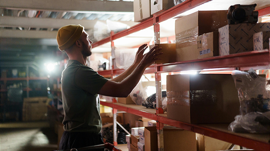 Warehouse worker in a cap checking boxes on red metal shelves in a lit room