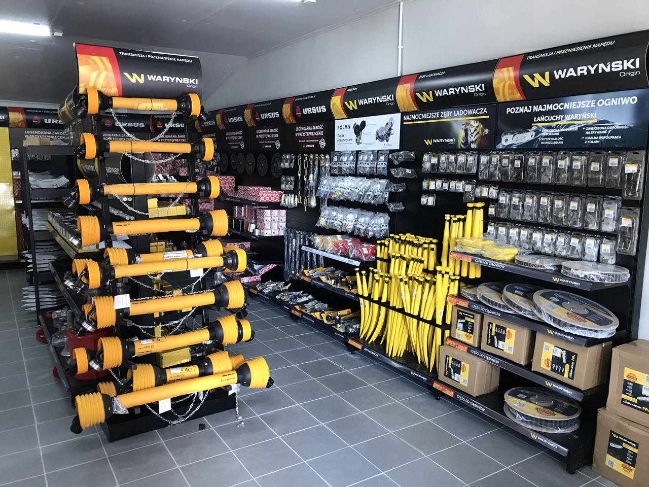 Store corner with Warynski products, showing yellow driveshafts on stand to the left and loader ribs and other tools on right. Warynski and Ursus brand advertising banners visible on walls