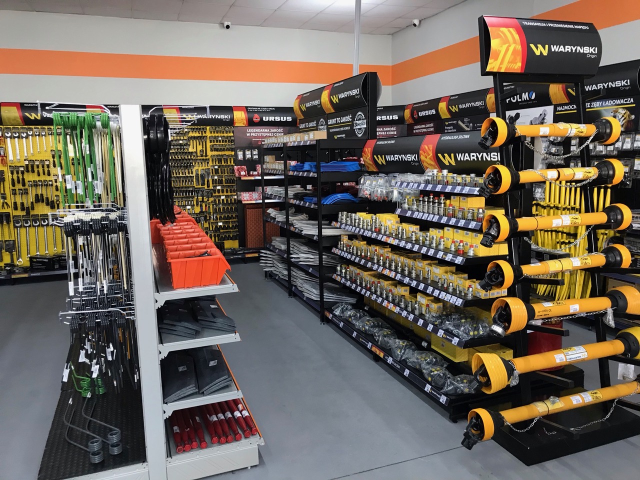 Store aisle with Warynski product display, showing various tools: green and red components on left, yellow driveshafts on right, and wall with tools and Warynski and Ursus advertising banners. Distinctive orange decorative strip visible on walls