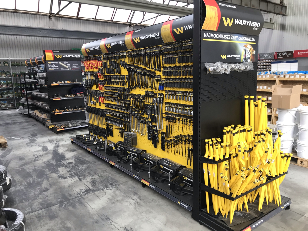 Warynski tools display in store, featuring black and yellow shelves with organized tools on perforated wall. Yellow crowbars and loader ribs visible on right, Warynski logo and branding above shelves